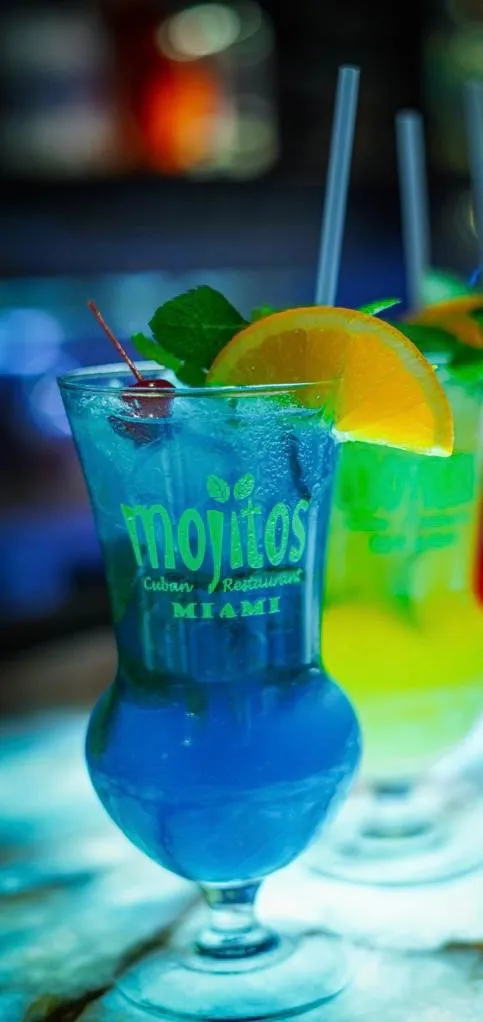 Primer plano de un mojito de curazao azul con ramita de menta, gajo de naranja y cereza junto a un cóctel amarillo en Mojitos Calle 8, restaurante cubano en Miami