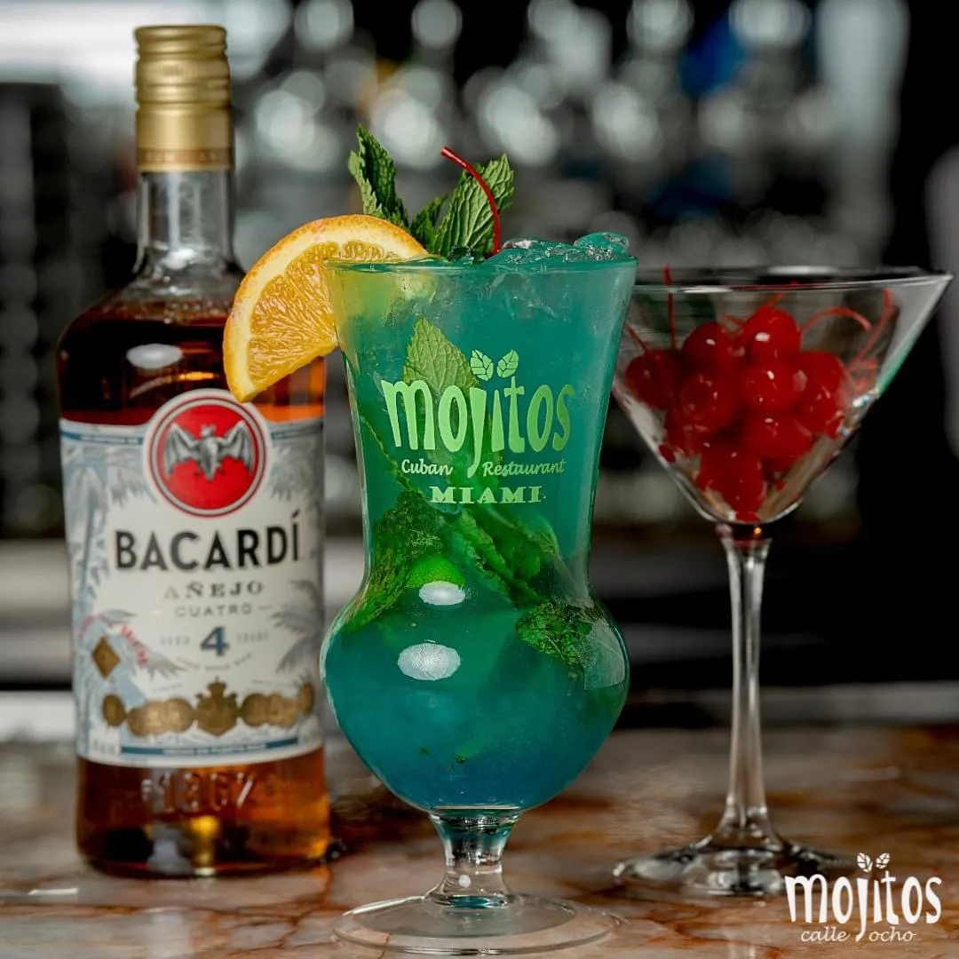 Mojito azul turquesa con menta y rodaja de naranja junto a una botella de ron Bacardi Añejo Cuatro y una copa martini con cerezas marrasquino en Mojitos Calle 8, restaurante cubano en Miami