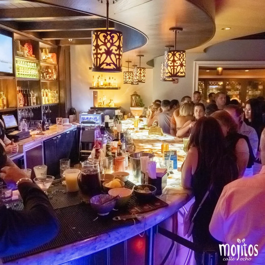 Crowded bar at night with guests drinking cocktails under ornate pendant lamps at Mojitos Calle 8, Cuban restaurant in Miami