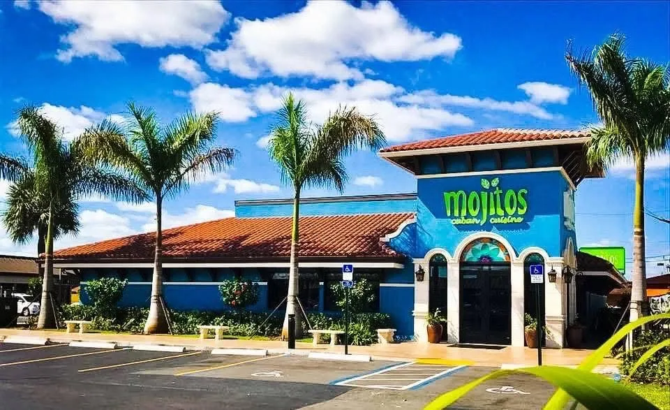 Daytime exterior of the iconic blue building with red tile roof, palm trees and parking lot, Mojitos Calle 8 on SW 8th Street, Miami