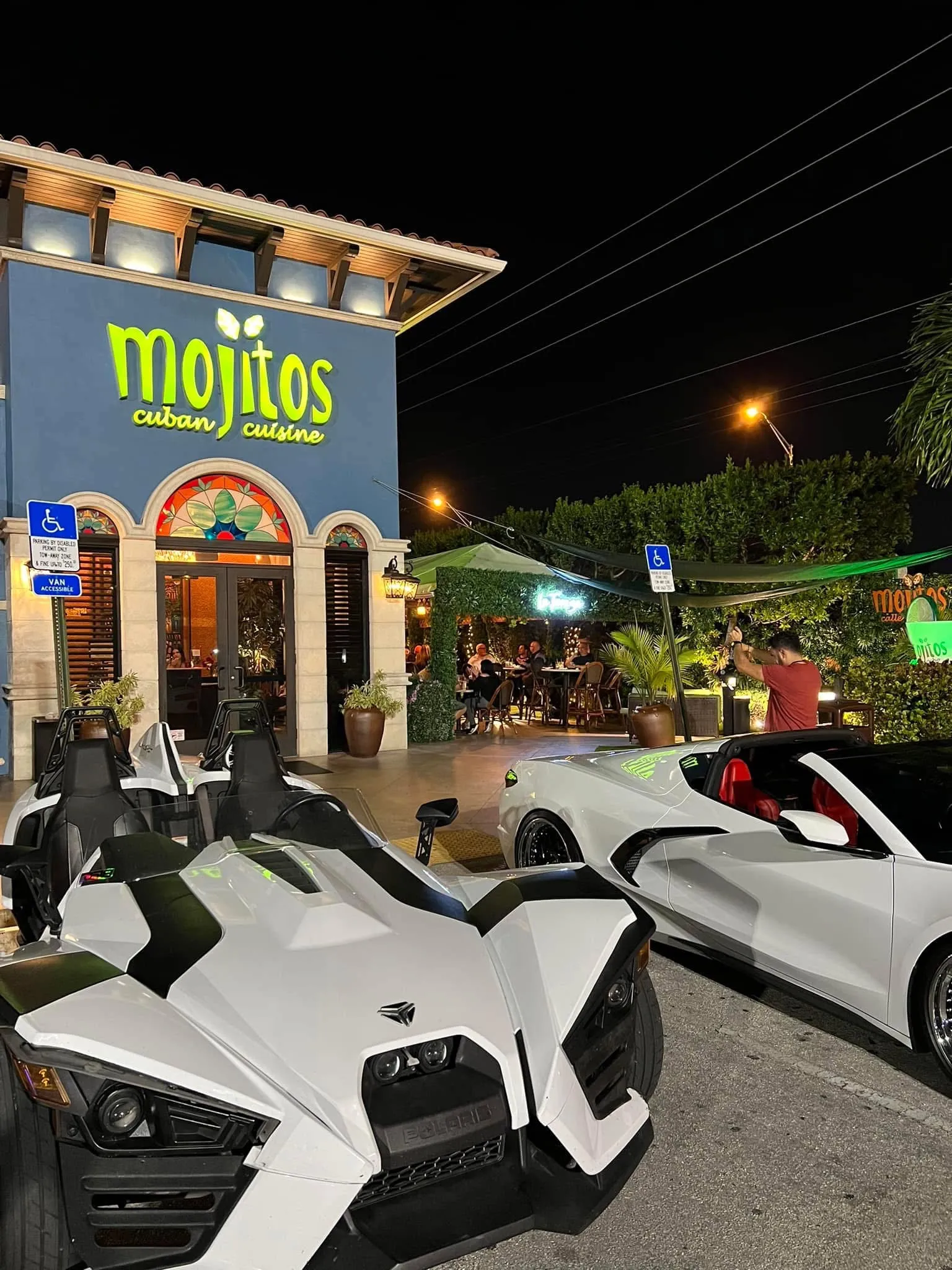 Sports cars and a Polaris Slingshot parked outside the blue building at night with the terrace busy behind, Mojitos Calle 8 Miami