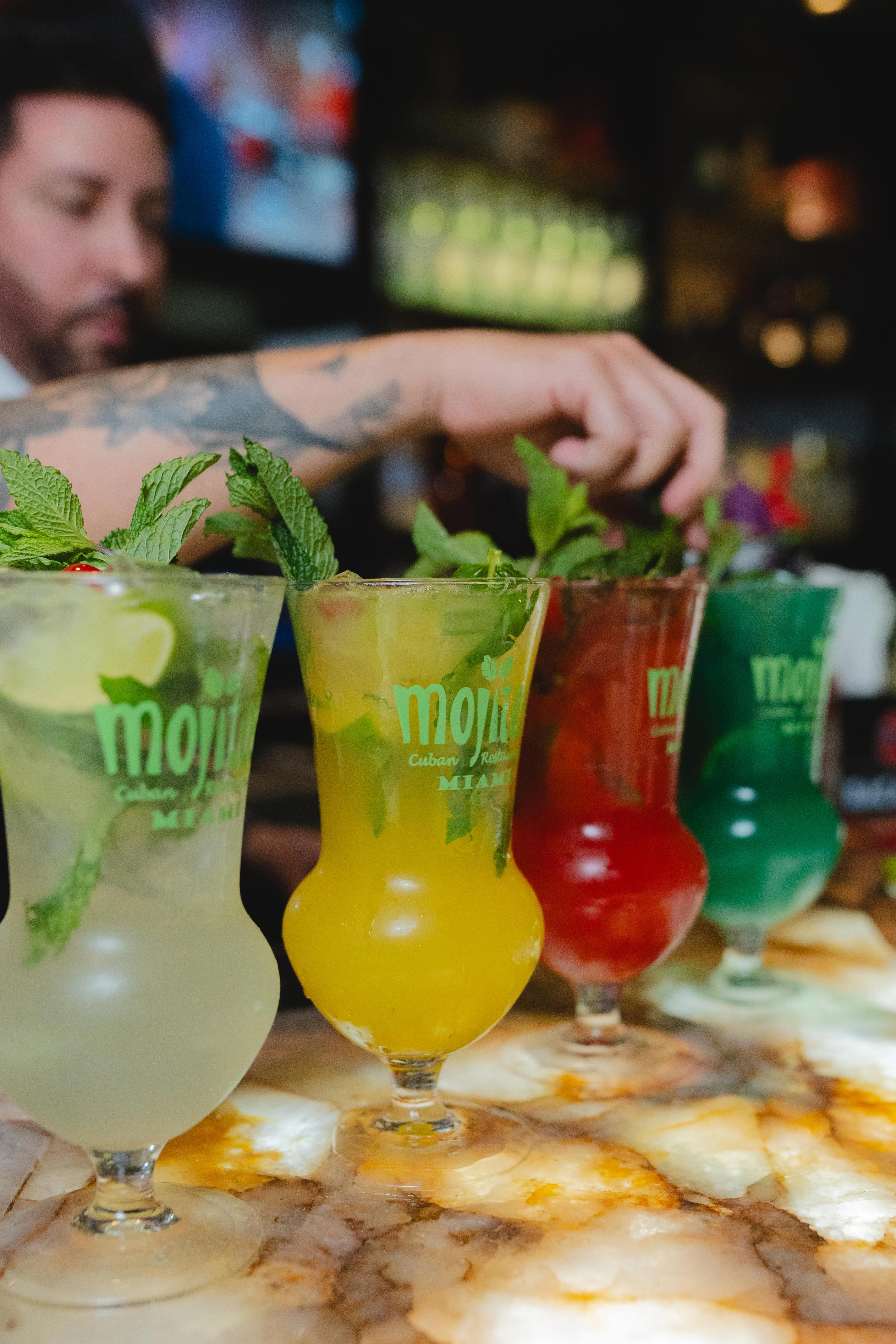 Fila de mojitos de sabores clásico, maracuyá, fresa y melón verde coronados con menta mientras el bartender los decora en Mojitos Calle 8, restaurante cubano en Miami