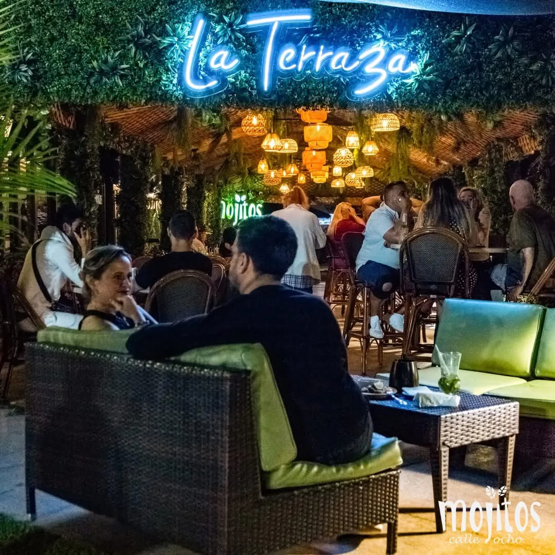 Guests relaxing on lounge sofas under the glowing La Terraza neon sign and lantern-lit pergola at Mojitos Calle 8, Miami