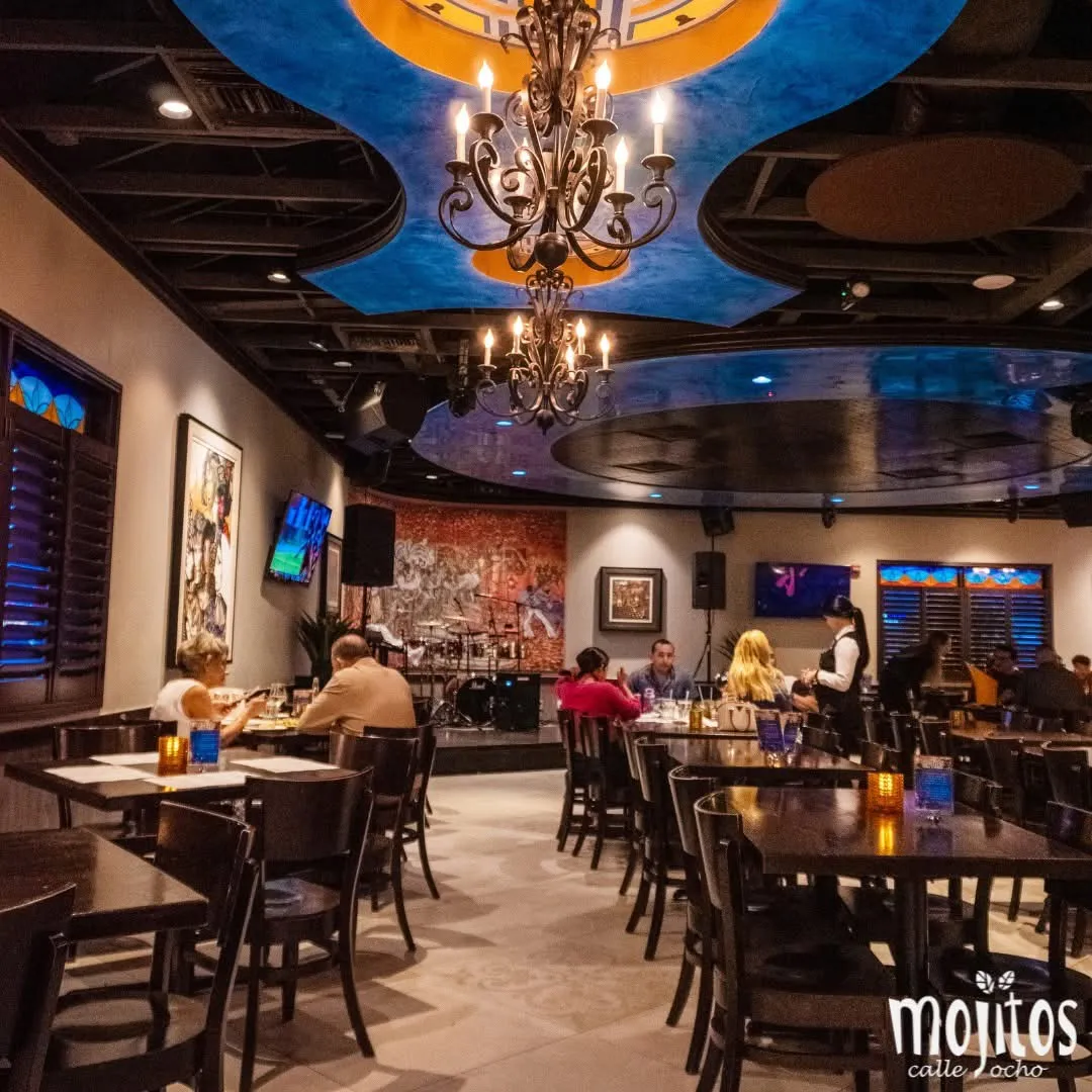 Main dining room with wrought-iron chandeliers, painted ceiling and the live music stage with drum kit at Mojitos Calle 8, Miami