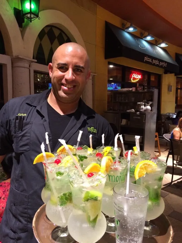 Mesero sonriente cargando una bandeja grande de mojitos decorados con rodajas de naranja y cerezas en la terraza de Mojitos Calle 8, restaurante cubano en Miami