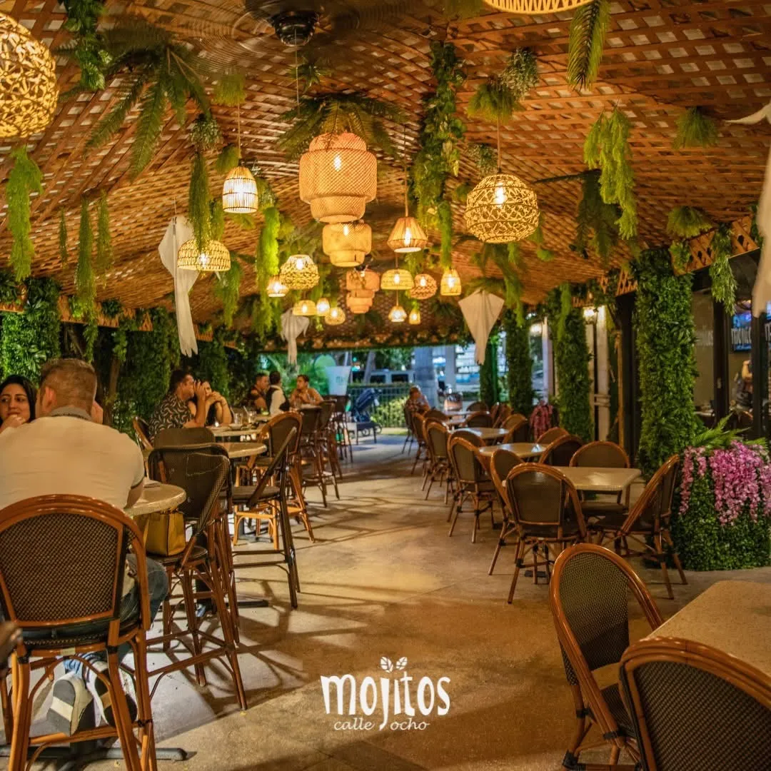 Evening on the covered terraza with diners seated beneath rattan pendant lights and hanging ferns at Mojitos Calle 8, Miami