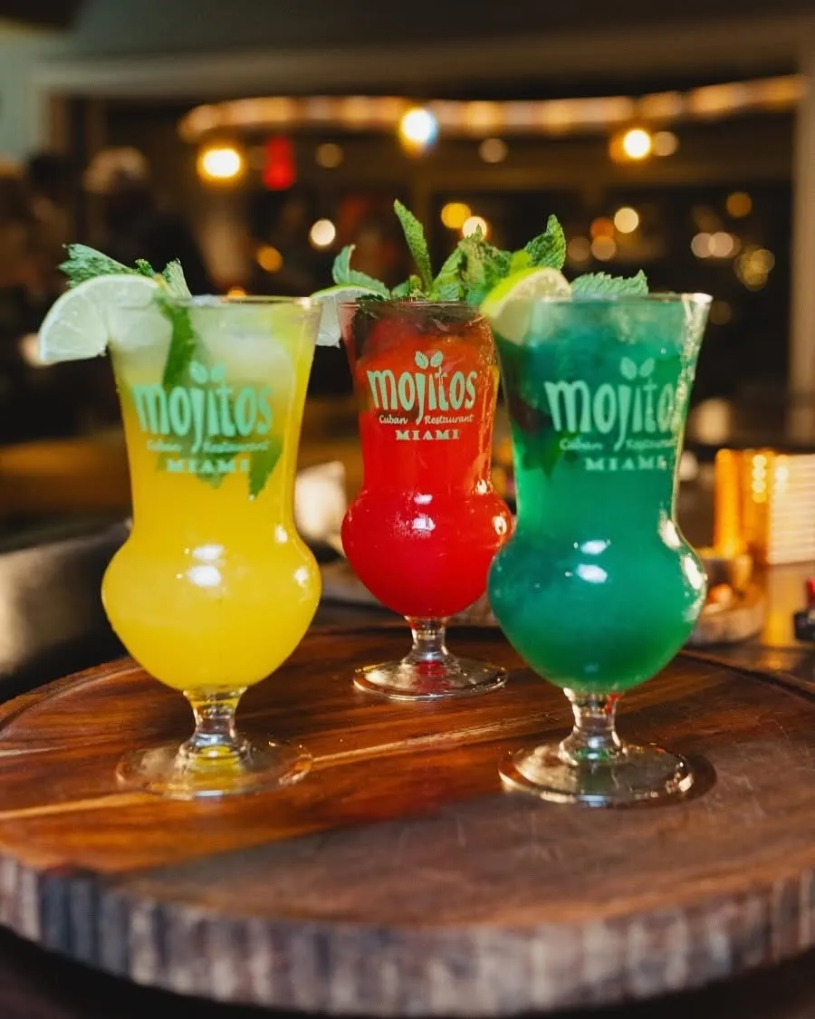 Tres mojitos de colores en amarillo mango, rojo fresa y verde curazao servidos sobre tabla redonda de madera con limón y menta en Mojitos Calle 8, restaurante cubano en Miami