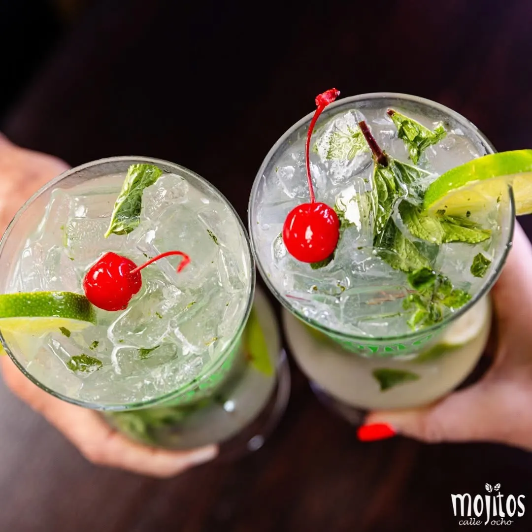 Vista cenital de dos manos brindando con mojitos clásicos llenos de hielo frapé, menta, limón y cerezas marrasquino en Mojitos Calle 8, restaurante cubano en Miami